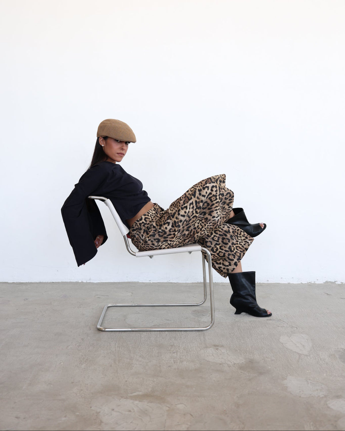 A model wearing a tan flat cap, black blazer, leopard-print skirt, and black peep-toe boots sits on a chrome cantilever chair against a white wall and concrete floor.