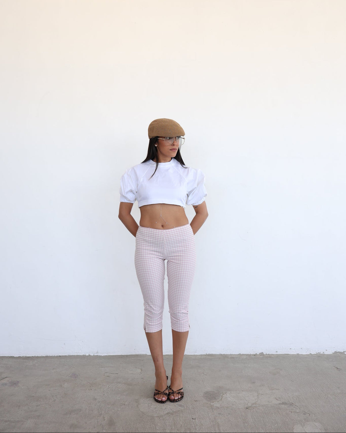 A woman stands against a plain white wall wearing a white cropped puff-sleeve top, light pink gingham capri pants, a tan beret, and black sandals.