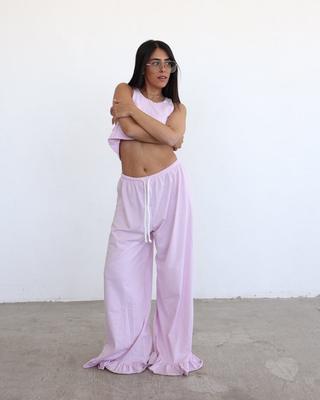 A woman wearing a light pink two-piece lounge set consisting of a sleeveless crop top and matching wide-leg pants, standing against a plain white wall and concrete floor.