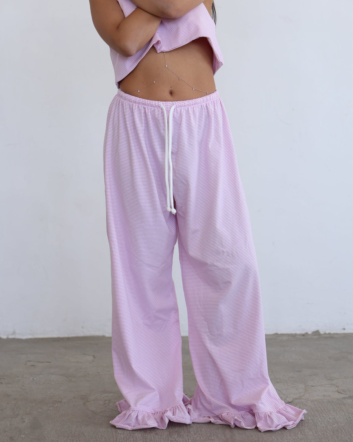 A model wearing light pink lounge pants with a white drawstring and a matching light pink cropped top, standing against a plain white wall and concrete floor.