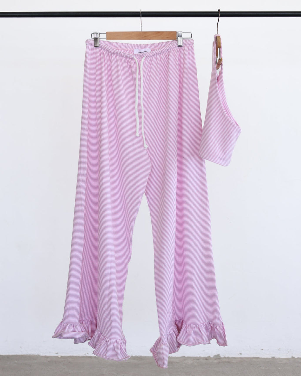 Light pink wide-leg pants with a drawstring waist and ruffled hems, shown hanging from a wooden hanger next to a matching sleeveless top on a black clothing rack against a white wall.